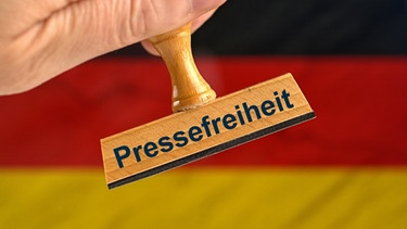 Ein symbolischer Holzstempel mit der Aufschrift "Pressefreiheit", gehalten von einer Hand im Anschnitt vor einer unscharfen Deutschland-Flagge im Hintergrund.  | Bild: picture alliance / SULUPRESS.DE | Torsten Sukrow