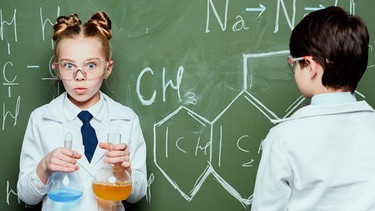 Zwei Kinder stehen an einer Tafel. Das Mädchen hält Reagenzgläser in der Hand, der Junge schreibt etwas an die Tafel. | Bild: colourbox.com