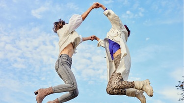Zwei Frauen halten sich die Hände und hüpfen in die Luft. Soziales Miteinander und Empathie stärkt Beziehungen.  | Bild: Wochit/Getty Images/Maki Nakamura