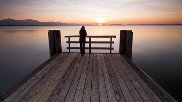 Frau steht alleine an einem Steg und betrachtet den Sonnenuntergang | Bild: picture alliance/imageBROKER