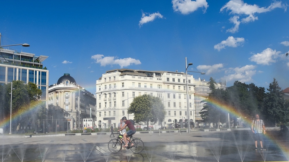 Ein Mann auf einem Fahrrad fährt auf einem Platz durch Wasser. Dabei bildet sich ein Regenbogen. Städte werden immer öfter zu Hitzeinseln. In Metropolen ist es oft heißer als auf dem Land.  | Bild: picture-alliance/dpa