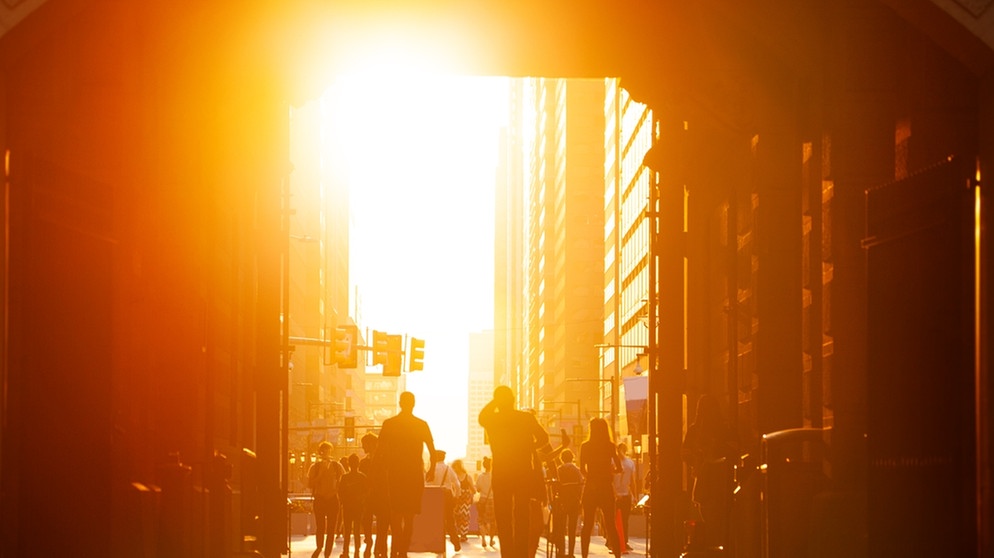 Mehrere Menschen gehen durch eine Innenstadt. Im Hintergrund sind die Sonne und eine Straße mit Autos zu sehen. Hitzetage in Städten nehmen deutlich zu. Pflanzen können gegen die hohen Temperaturen helfen. | Bild: colourbox.com