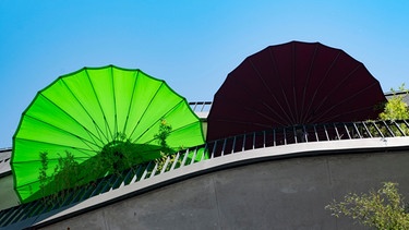 Zwei Sonnenschirme in Grün und Schwarz sind auf einem Balkon aus Beton zu sehen. In Städten nehmen die Hitzewellen zu. Pflanzen und eine nachhaltige Bauweise können gegen Hitze in Städten helfen. | Bild: picture-alliance/dpa