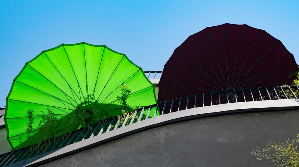 Zwei Sonnenschirme in Grün und Schwarz sind auf einem Balkon aus Beton zu sehen. In Städten nehmen die Hitzewellen zu. Pflanzen und eine nachhaltige Bauweise können gegen Hitze in Städten helfen. | Bild: picture-alliance/dpa