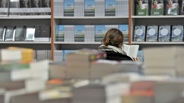 Vordergrund Bücherstapel, Frau liest vor Bücherwand | Bild: picture-alliance/dpa