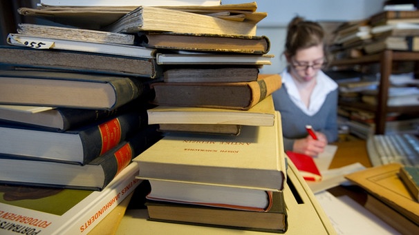Bücherstapel im Vordergrund, Frau schreibend dahinter | Bild: picture-alliance/dpa
