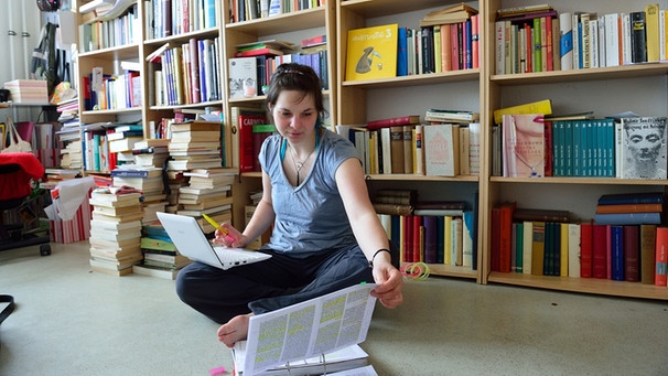 Studentin mit Laptop auf Boden sitzend, blättert im Ordner, Hintergrund Bücherregal | Bild: picture-alliance/dpa