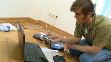 Eric kniet vor einem Laptop am Boden mit seinem Handy in der Hand, um ihn liegen Gebrauchsanweisungen. | Bild: BR