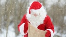 Ein Weihnachtsmann steht im Schnee und schaut in seinen Sack nach Geschenken. | Bild: colourbox.com