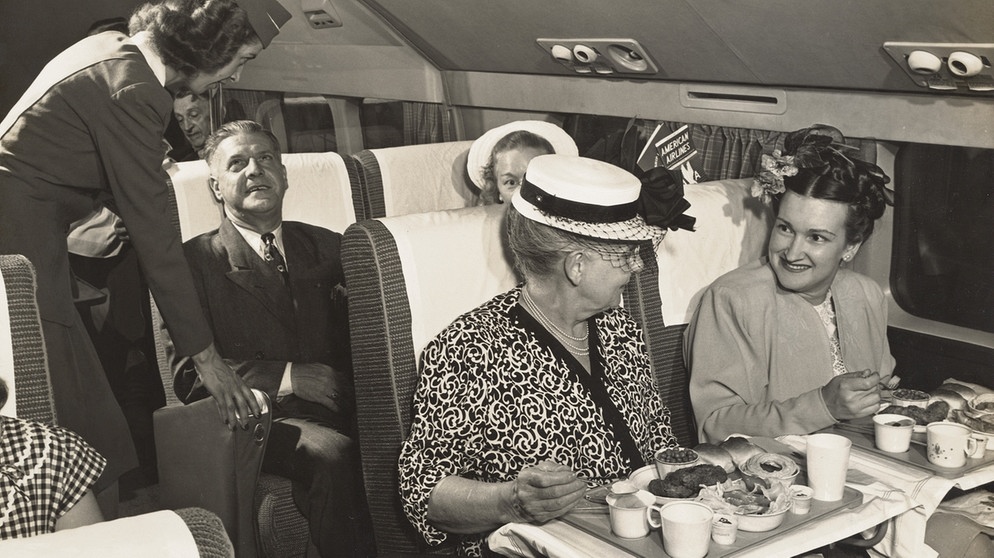 Frauen mit Hüten essen im Jahr 1947 in der Ersten Klasse einer American Airlines Dc-6, dahinter spricht eine Stewardess mti einem weiteren Passagier. Seit 100 Jahren servieren Airlines ihren Passagieren Essen im Flugzeug. | Bild: picture alliance/Mary Evans Picture Library