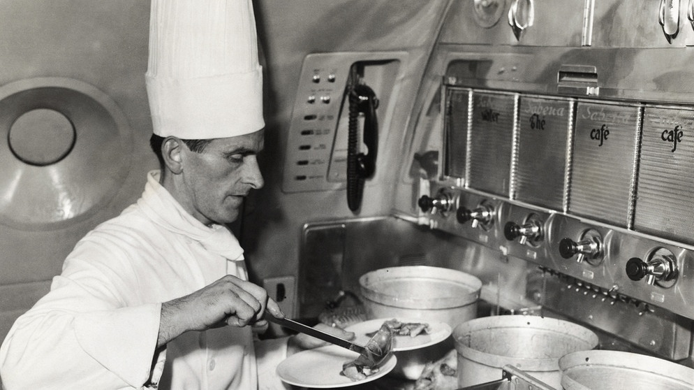 Ein Koch mit hoher Kochmütze richtet das Essen auf einem Teller an, im Jahr 1940 gab es das an Bord einer Sabena DC-6. Seit 100 Jahren servieren Airlines ihren Passagieren Essen im Flugzeug. | Bild: picture alliance/Mary Evans Picture Library