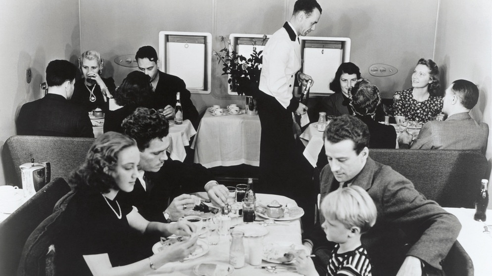Drei Erwachsene und ein Kind sitzen im Jahr 1930 um einen Tisch an Bord einer Martin M-130 China Clipper und essen. Seit 100 Jahren servieren Airlines ihren Passagieren Essen im Flugzeug. | Bild: picture alliance/Mary Evans Picture Library