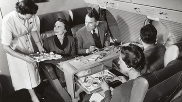 Eine Stewardess serviert im Jahr 1960  in der Erste Klasse Kabine den Passagieren Essen und Drinks. Seit 100 Jahre servieren Fluggesellschaften ihren Passagieren Essen. | Bild: picture alliance/Mary Evans Picture Library