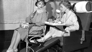 17.01.1957, Großbritannien, London: Janice van Ayre (l) hat auf einem Flugzeugsitz von 1928 Platz genommen, auf dem Schoß eine Schachtel mit Essen. Etwas bequemer hat es Anne Grierson (r) mit ihrem bequemen "Slumberette" Sitz der Fluggesellschaft B.O.A.C., der auch zum Schlafen genutzt werden kann. Sie bekommt das Essen auf einem Tablett serviert. | Bild: dpa-Bildfunk/UPI