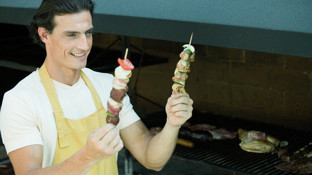 Ein Mann, der vor einem Grill steht, hält zwei Fleischspieße in der Hand | Bild: colourbox.com