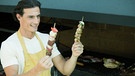 Ein Mann, der vor einem Grill steht, hält zwei Fleischspieße in der Hand | Bild: colourbox.com