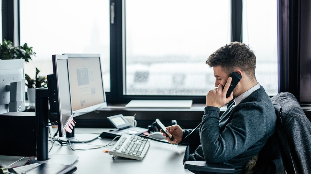 Ein Angestellter telefoniert am Festnetz und bedient gleichzeitig sein Mobiltelefon. | Bild: BR/Johanna Schlüter