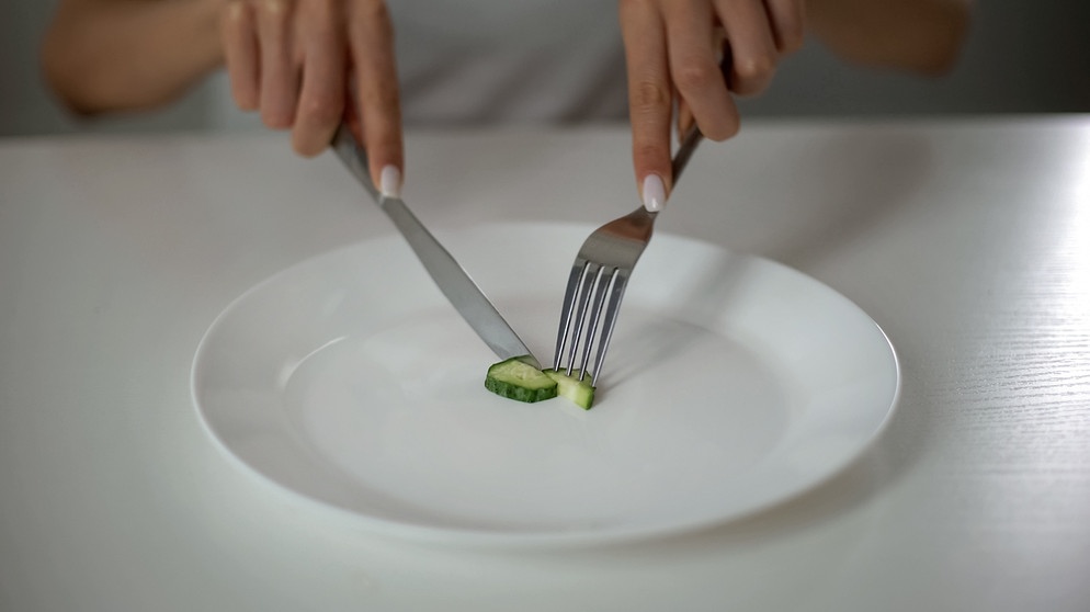 Eine Frau (es sind nur die Hände zu sehen) sitzt vor einem weißen Teller, auf dem eine Scheibe Gurke zu sehen ist, die sie mit Messer und Gabel zerteilt. | Bild: Colourbox.de