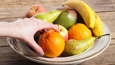 Eine Hand greift in eine Schüssel mit Obst wie Bananen, Orangen, Birnen, Äpfel und Mango. | Bild: Colourbox.de