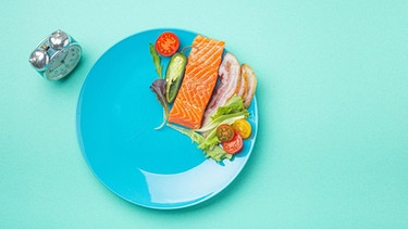 Auf einem türkisen Untergrund steht ein Wecker und ein blauer Teller. Darauf einige Lebensmittel, die von 12 Uhr bis 16 Uhr angeordnet sind: Lachs, Bacon, Salat. | Bild: NDR