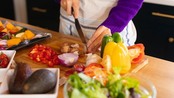 Eine blonde Frau steht mit Schürze über den Anziehsachen in einer Küche, schneidet Gemüse klein, das sie mit einem großen Messer von einem Holzbrett gibt. Im Hintergrund diverse gesunde Gemüsesorten. | Bild: picture alliance / Zoonar | Channel Partners