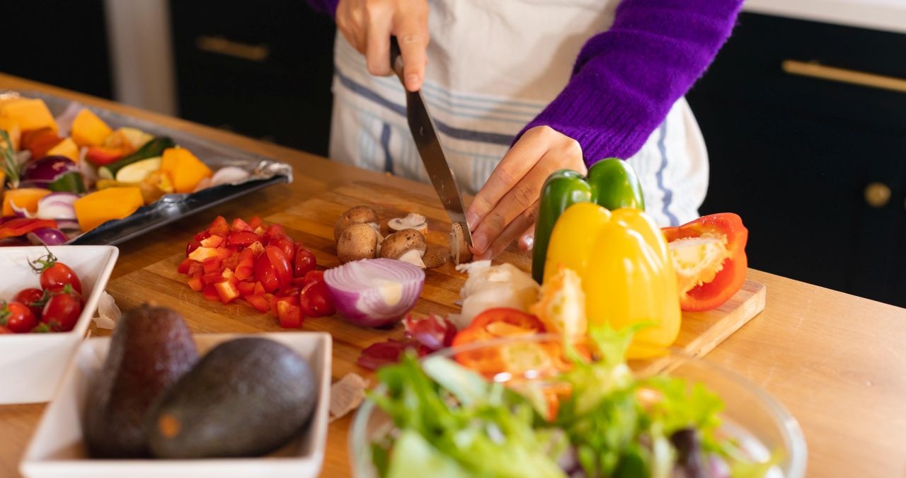 Eine blonde Frau steht mit Schürze über den Anziehsachen in einer Küche, schneidet Gemüse klein, das sie mit einem großen Messer von einem Holzbrett gibt. Im Hintergrund diverse gesunde Gemüsesorten. | Bild: picture alliance / Zoonar | Channel Partners