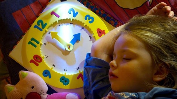 Ein Mädchen schläft in einem Bett, auf dem Kopfkissen liegt eine Kinder-Uhr | Bild: picture-alliance/dpa