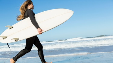 Eine junge Frau läuft mit ihrem Surfbrett über den Strand zum Wasser. Können wir Menschen bald schon viel länger gesund leben? Wir erklären euch aussichtsreiche wissenschaftlichen Entdeckungen der Alternsforschung für ein gesundes Altern und ein längeres Leben. | Bild: colourbox.com/Ikostudio
