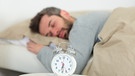 Ein Mann schläft im Hintergrund eines Weckers. Warum schlafen wir eigentlich? Wenn wir schlafen, verarbeitet unser Gehirn Erlebtes und sortiert Unwichtiges aus. | Bild: colourbox.com