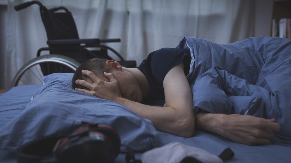 Mann mit MECFS liegt im Bett und schützt seine Augen. Im Vordergrund eine Schlafmaske und Noise-Cancelling-Kopfhörer, hinter dem Bett ein Rollstuhl. | Bild: Lea Aring