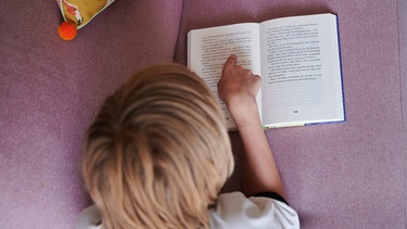 Ein Junge liest ein Buch auf dem Sofa. Gut lesen zu können, ist wichtig für das erfolgreiche Lernen. Wie ihr Kinder beim Lesenlernen unterstützen könnt und warum Vorlesen eine besondere Rolle spielt. | Bild: picture alliance/dpa/Annette Riedl 