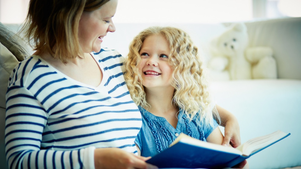 Eine Mutter liest mit ihrer Tochter auf dem Sofa ein Buch. Lesenlernen ist eine wichtige Voraussetzung dafür, dass Kinder erfolgreich lernen können. Wir erklären euch, wie ihr Kinder dabei unterstützen und fördern könnt. | Bild: colourbox.com/ Dmitrii Shironosov