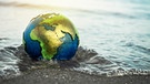 Eine Weltkugel wird an einem Strand von Wasser überspült. Durch die globale Erwärmung infolge des Klimawandels steigt der Meeresspiegel. | Bild: picture-alliance/dpa