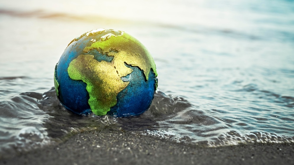Eine Weltkugel wird an einem Strand von Wasser überspült. Durch die globale Erwärmung infolge des Klimawandels steigt der Meeresspiegel. | Bild: picture-alliance/dpa