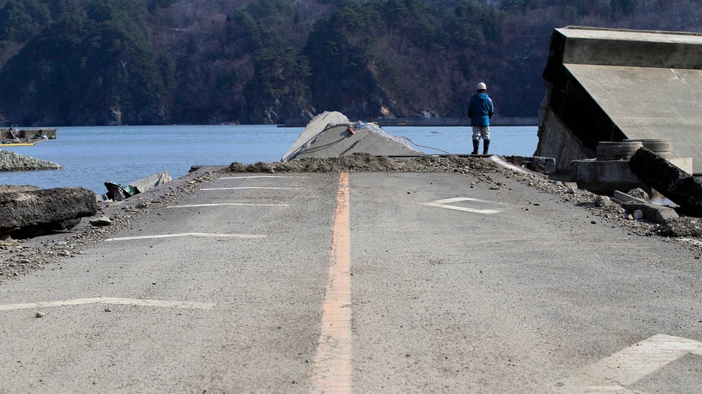 Straßenarbeiter steht an einer zerstörten Straße in Kamaishi in der Präfektur Iwate  im Nordosten Japans nach dem schweren Erdbeben und Tsunami vom 11. März 2011. Starke seismische Aktivitäten und Verschiebungen tektonischer Platten können riesige Flutwellen, japanisch: Tsunamis auslösen. | Bild: picture alliance / dpa | Stephen Morrison