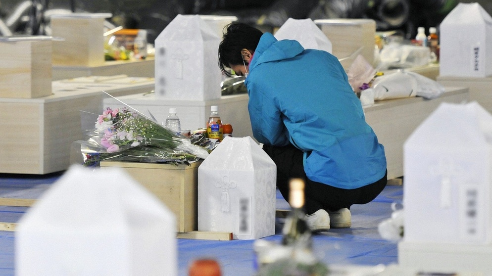 Ein Mann hockt neben einem Sarg in einem Gedenkraum in Soma, Präfektur Fukushima am 27. März 2011 nach dem verheerenden Tsunami und Erdbeben im Nordosten Japans. Die Naturgewalten beschädigten auch das Atomkraftwerk Fukushima: eine nukleare Katastrophe. | Bild: picture-alliance/dpa