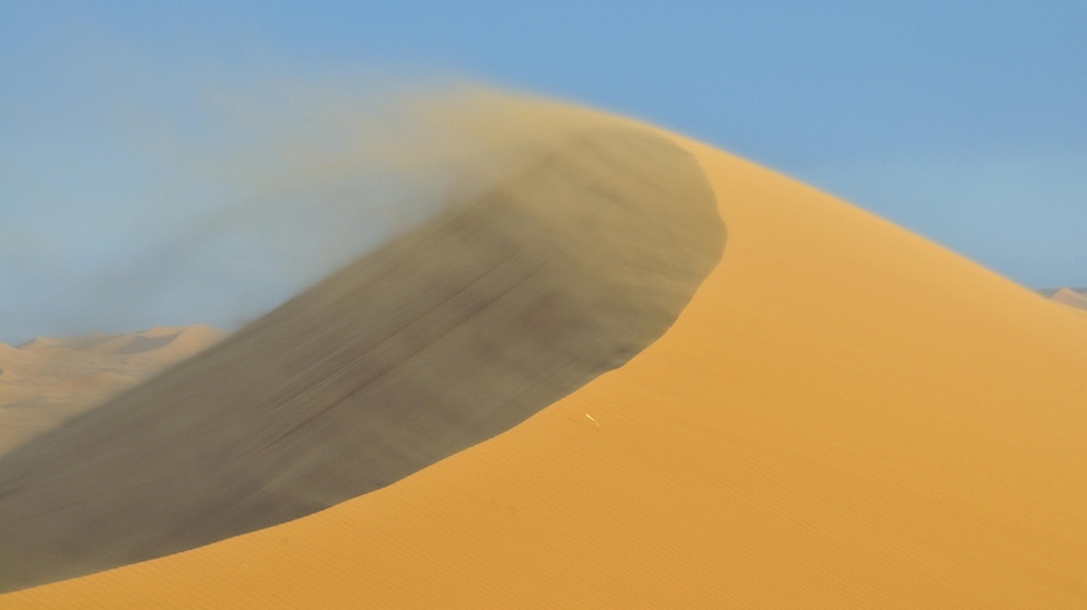 Ein Sandsturm in der Wüste Erg Chebbi in Marokko bläst losen Sand vom Kamm einer Sanddüne. Sand, Wind und Schwerkraft - das sind die drei Hauptfaktoren für die Bildung von beeindruckend geformten Sanddünen in Wüsten. | Bild: picture-alliance/imageBROKER