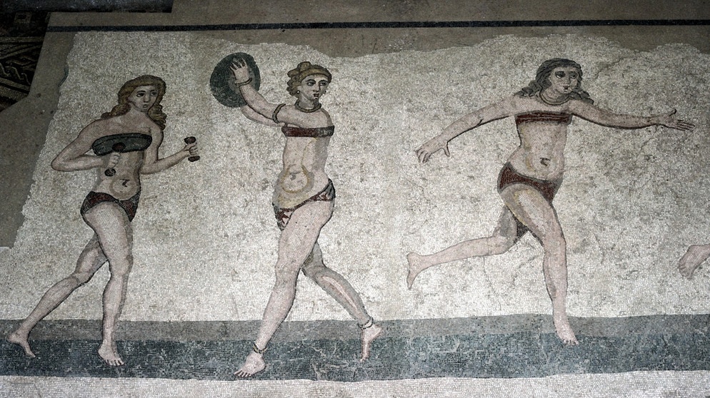 Ein römisches Mosaik aus dem 4. Jahrhundert. n. Chr. zeigt die sogenannten "Bikinimädchen" in der Villa Romana del Casale, Sizilien. Viele Menschen treibt die Angst um, in ihrer Freizeit etwas zu verpassen. Wir haben Tipps gegen FOMO und Freizeitstress, damit ihr euch in eurer freien Zeit wirklich erholt. | Bild: picture alliance / Artcolor 