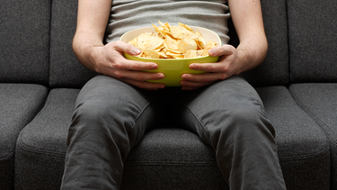 Mann sitzt mit einer Schüssel Chips auf dem Sofa. Routinen und Rituale entlasten unser Gehirn bei seinen täglichen Aufgaben. Aber nicht alle Gewohnheiten sind nützlich. Wie wir schlechte Angewohnheiten loswerden und unser Verhalten zum Positiven verändern können. | Bild: colourbox.com