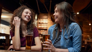 Zwei glückliche junge Frauen trinken gemeinsam ein Bier am Tisch. Was ist Glück? Wann fühlt man es und wie? Und kann man dem Glücksgefühl auch auf die Sprünge helfen? Wir haben für euch Infos und Tipps zum Glücklichsein gesammelt. | Bild: picture alliance / Westend61 | gpointstudio