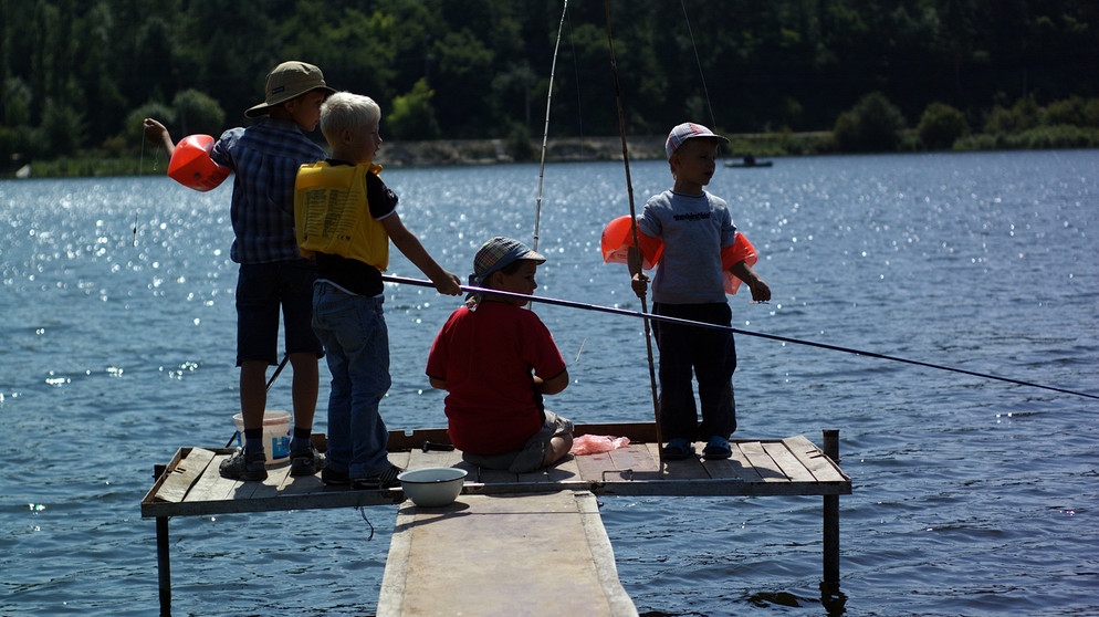 Kinder sitzen auf einem Steg am See und angeln. Eltern tun gut daran, ihre Kinder auch mal eigene Erfahrungen sammeln zu lassen. Das stärkt das Selbstvertrauen.  | Bild: colourbox.com