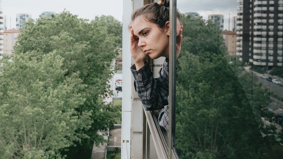 Eine junge Frau lehnt sich besorgt aus dem Fenster eines Hochhauses | Bild: colourbox.com