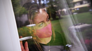 Frau mit Maske blickt aus dem Fenster.  | Bild: picture-alliance/dpa