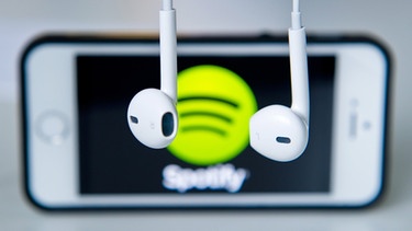 Kopfhörer hängen vor einem Smartphone, auf dem das Logo vom Musik-Streaming-Dienst Spotify angezeigt wird. | Bild: dpa-Bildfunk/Daniel Bockwoldt