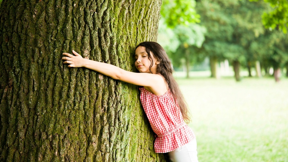 Mädchen umarmt lächelnd einen Baum. Umarmt euch öfter - nicht nur zum Weltknuddeltag am 21. Januar! Warum Nähe ein Grundbedürfnis und Kuscheln gesund ist. | Bild: picture alliance / imageBROKER | uwe umstätter
