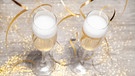 Das Entfernen des Sektkorkens an Silvester oder Neujahr für den perfekten Knall ist gar nicht so einfach. Im Bild: Zwei Champagnergläser mit weißem Schaum stehen auf einem Tisch. | Bild: picture-alliance/dpa