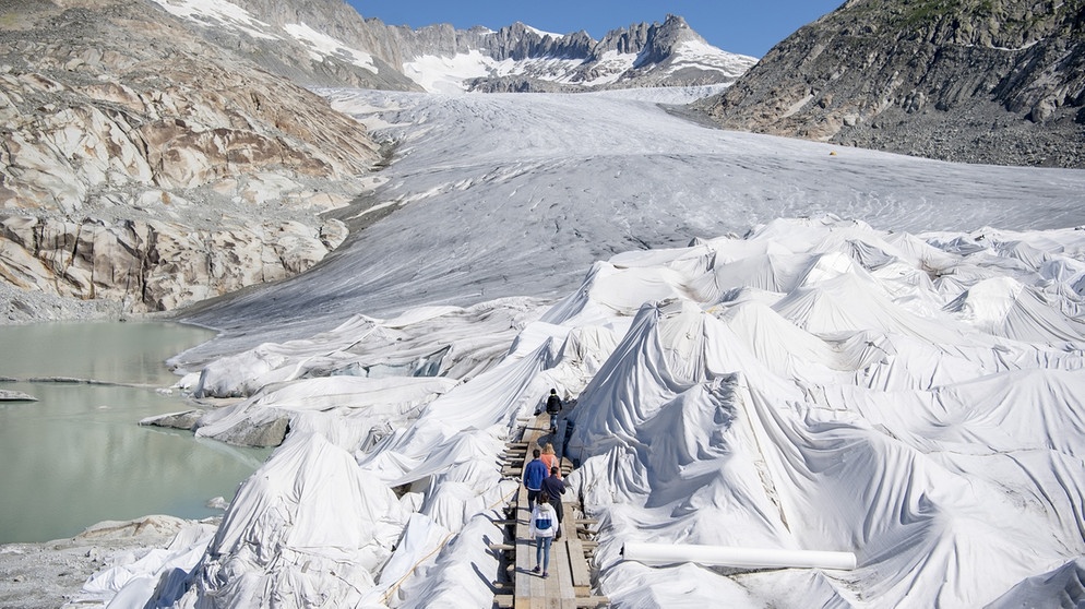 Touristen besuchen die mit Planen abgedeckte Eisgrotte am Gletscherende des Rhonegletscher oberhalb von Gletsch am Furkapass.  | Bild: picture-alliance/KEYSTONE | URS FLUEELER