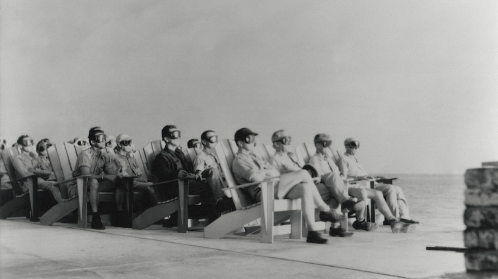 Schutzbrillen, eine Art Strandstuhl und aus sicherer Entfernung: VIPs beobachten 1951 den ersten Test einer Detonation im Rahmen der Operation Greenhouse. Mit dieser Testserie probierten die USA aus, wie später eine Wasserstoffbombe funktionieren könnte.  | Bild: picture alliance / Everett Collection | -