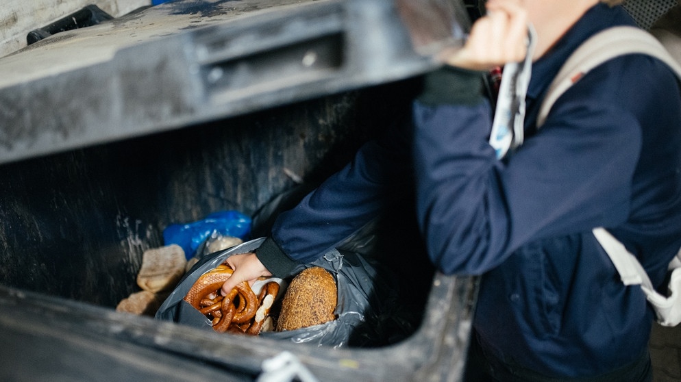 Eine Frau fischt Lebensmittel aus einer Mülltonne eines Supermarkts. Fast 60 Kilogramm Lebensmittel wirft jeder Deutsche pro Jahr in den Müll. Noch genießbare Lebensmittel aus der Tonne eines Supermarkts zu fischen ("Containern") ist nicht legal. Es gibt andere Möglichkeiten, Essen vor der Tonne zu retten. | Bild: BR/Julia Müller