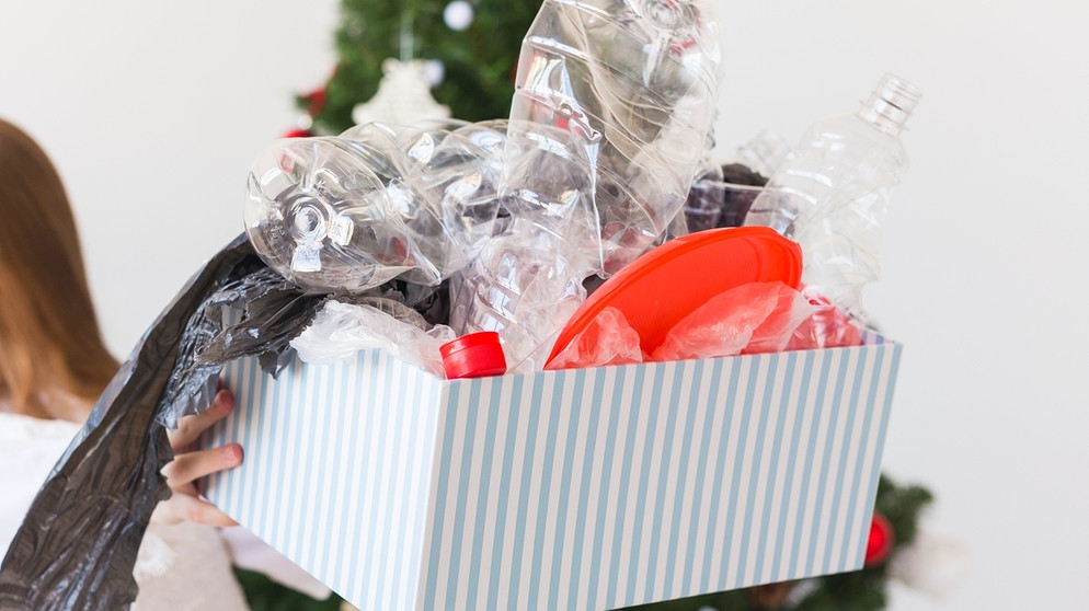 Ein Mädchen vor einem Weihnachtsbaum hält ein Paket voller Plastikmüll. Schätzungen zufolge produzieren wir 20 Prozent mehr Müll an Weihnachten. Weihnachtsbaum, Plätzchen, Braten und Geschenkpapier sind oft wenig umweltfreundlich. Wie ihr mit einfachen Tipps und Tricks ressourcenschonende und trotzdem schöne Weihnachten feiern könnt. | Bild: colourbox.com